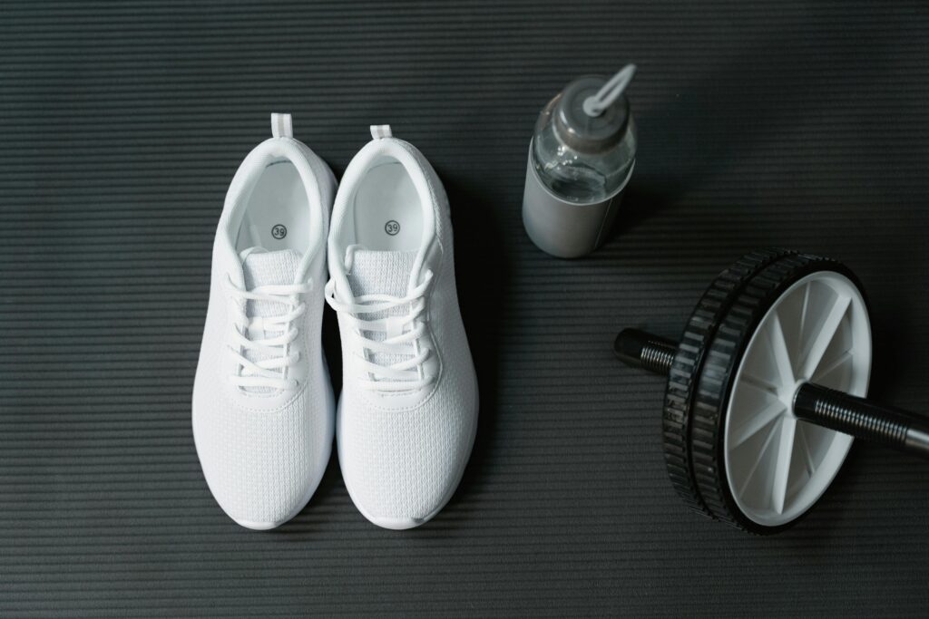 Flat lay of white sneakers, ab roller, and water bottle on black mat for fitness motivation.